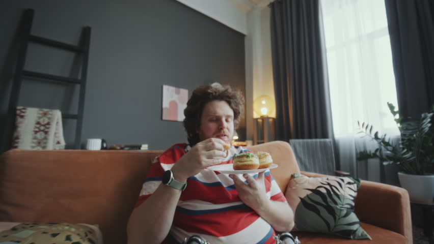 Young chubby man eating delicious donuts and enjoying their taste on couch in living room while watching TV at home. Whip zoom shot