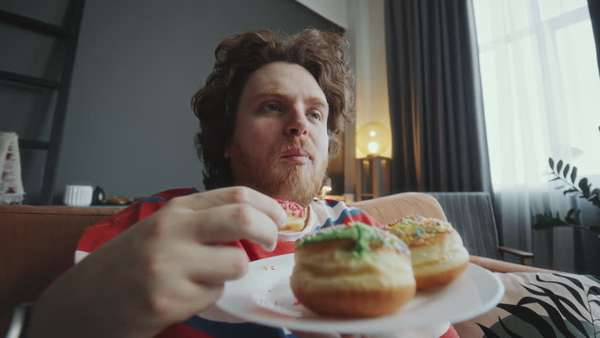 Young chubby man eating delicious donuts and enjoying their taste on couch in living room while watching TV at home. Whip zoom shot