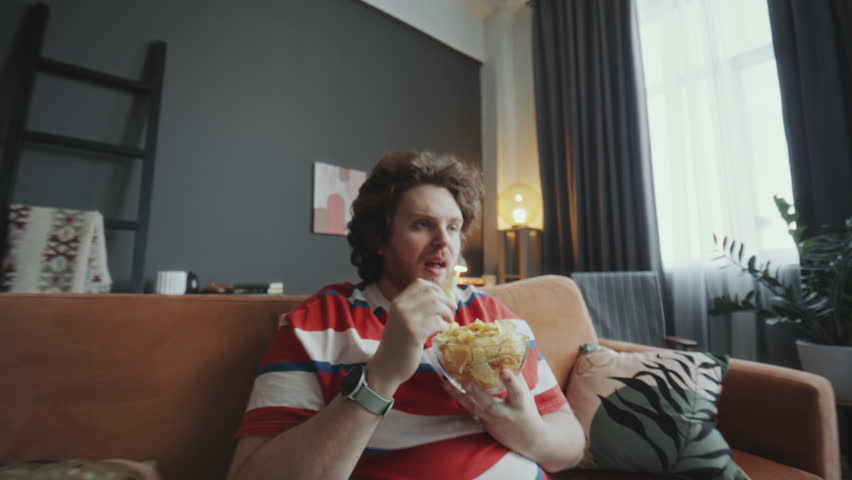 Young obese man relaxing on couch, eating potato chips from bowl and watching TV during day at home. Whip zoom shot