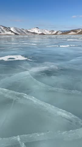 Vertical video of winter landscape of frozen Baikal lake on cold sunny day in February. Snow-capped hills frame iced bay with blue ice surface with cracks in mouth of Anga River