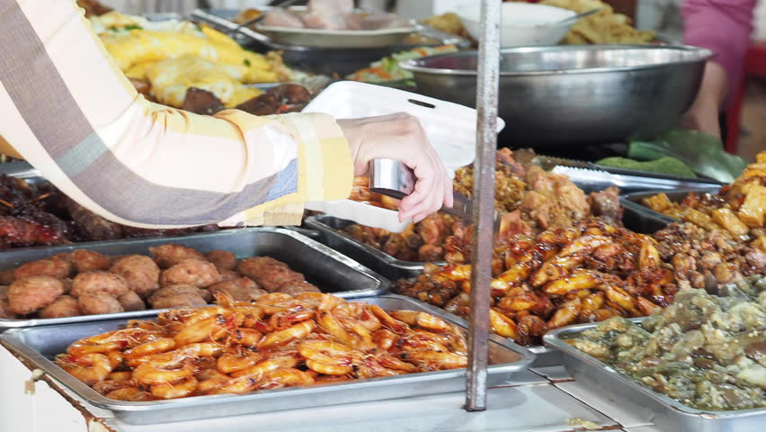 A woman uses tongs to take grilled food from metal buckets at a street eatery, placing it into a white basket for takeout at an Asian market.