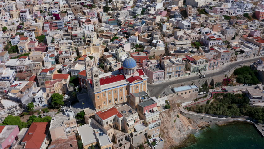 Experience a breathtaking aerial point-of-view shot of the Church of Agios Nikolaos at Asteria Beach, Syros Island, Greece. Marvel at its bright red roof and blue dome set within a cinematic cityscape