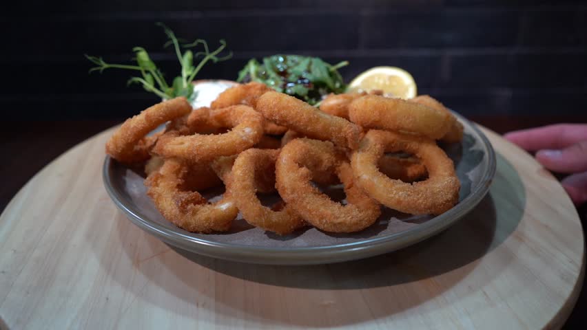 A plate of fried onion rings with sauce, lemon and green vegetables on a wooden spinning base