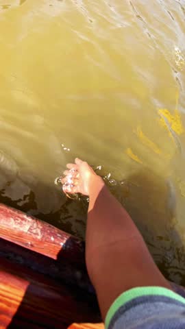 A person’s hand reaches out to touch the water while riding a wooden boat. The sunlight creates a warm atmosphere, highlighting the gentle movement of the water.