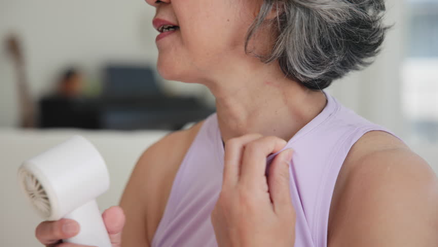 Asia adult people middle age old woman sit at sofa home feel hot flash fever use fan waving hand blowing wind face elder health care summer heat aging issue. Sad pain sweat tired stroke suffer stress.