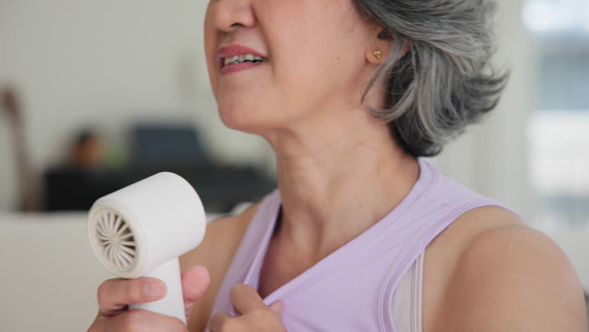 Asia adult people middle age old woman sit at sofa home feel hot flash fever use fan waving hand blowing wind face elder health care summer heat aging issue. Sad pain sweat tired stroke suffer stress.
