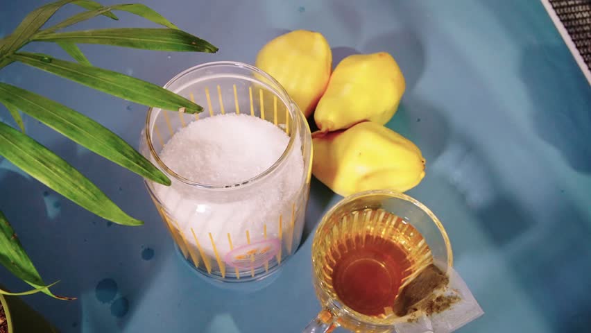 On the table are sugarv and an empty tea cup with a spoon. It is filled with tea leaves from a teapot. After that, the man's hand puts two pieces of sugar in the cup.