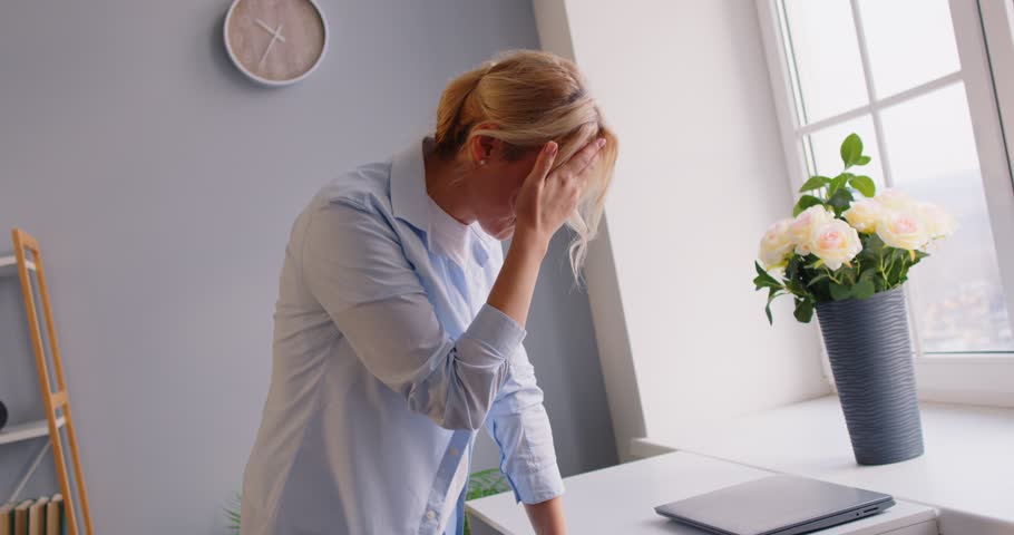 Tired blonde woman standing near window, touching forehead with hand, feeling bad, suffering from dizziness. Sick girl having vertigo, feeling exhausted, overheated or unwell, losing consciousness.