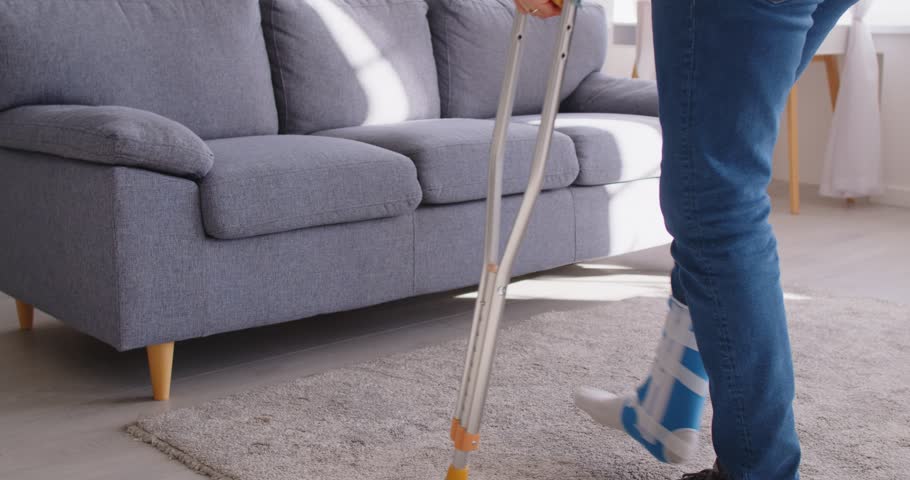 Rehabilitation after lower limb injury. Man wearing support brace and fracture fixator on broken leg and walking with crutches. Person sits on sofa and rubs his broken leg, feeling discomfort. 