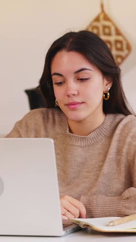 Vertical HD video of young happy woman working on laptop in the kitchen at home while drinking coffee in the morning. Female student learning online. Education or business concept