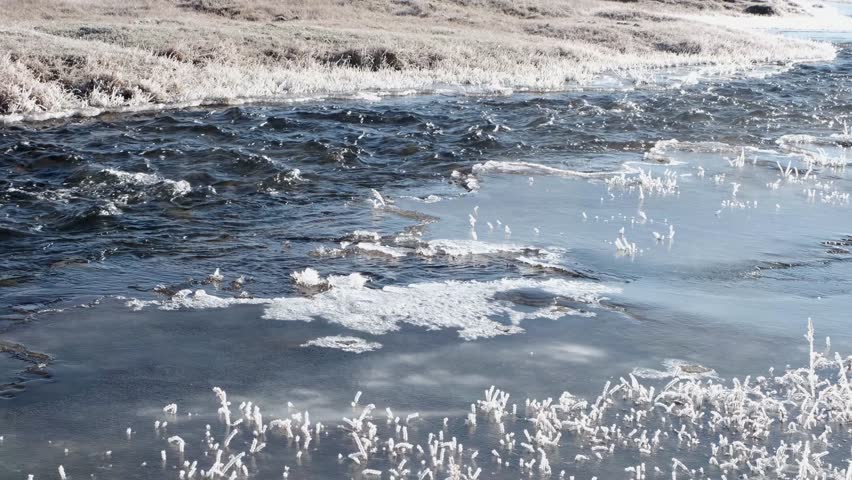 Fast-moving stream of narrow river runs along frozen banks on sunny day. Landscape of autumn nature with melting ice islands and frosted grass