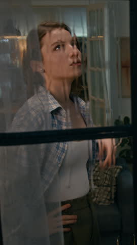 Medium vertical shot, watched through window from street, of young Caucasian couple fighting in living room on rainy night, screaming abuse and accusations at each other, gesticulating, shouting