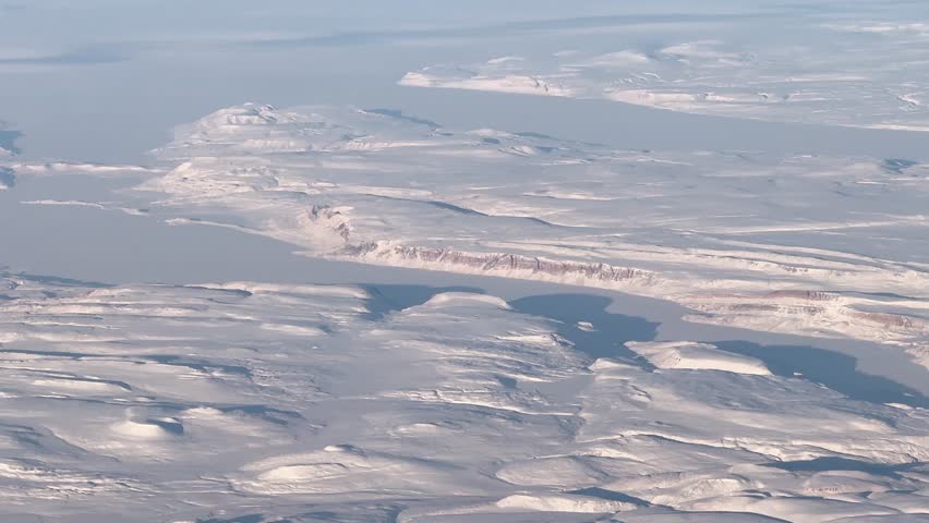 aerial video footage, flying across Greenland, approaching west coast in winter with a clear blue sky and white winter landscape below. Mountains covered with snow and ocean deeply frozen - 4k footage