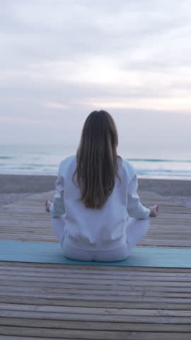 View from the back, brunette woman doing yoga on the beach by the sea. Vertical video.