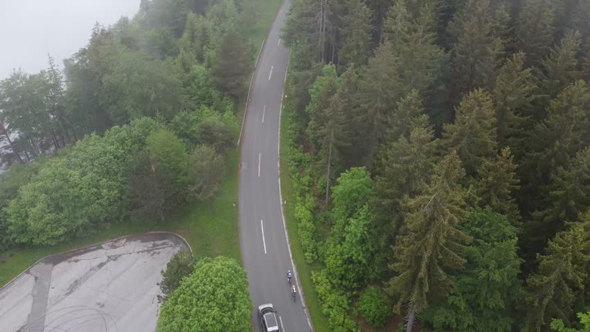Aerial drone view of road cyclists riding uphill towards dobratsch on villacher alpenpanorama road. Cloudy rainy weather in the mountain, green trees