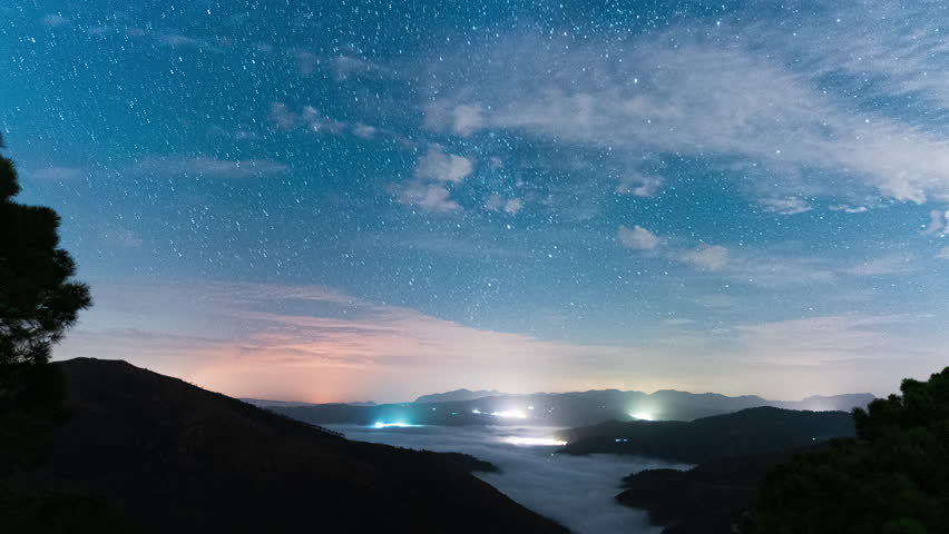 Milky Way galaxy spins above landscape of Spain with city lights, time lapse