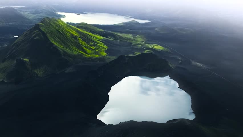 Aerial drone footage of a lake and green moss and grass covered steep tall mountains during a cloudy day in Greenland or Iceland. The lakes are reflective and white during cloudy day with mist and fog