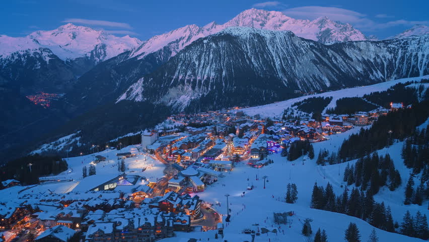Experience the stunning aerial view of nightlit Courchevel, France, with majestic snowcovered mountains above and twinkling village lights below, creating a magical and enchanting scene