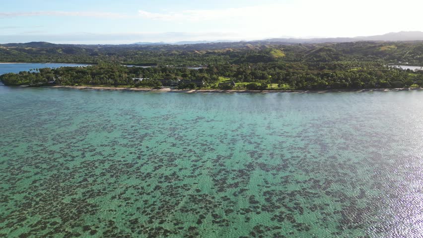 A drone view of coral reef seen under the water surface with a greenery-covered shoreline next to the Shangri-La Yanuca Island in Fiji, Oceania