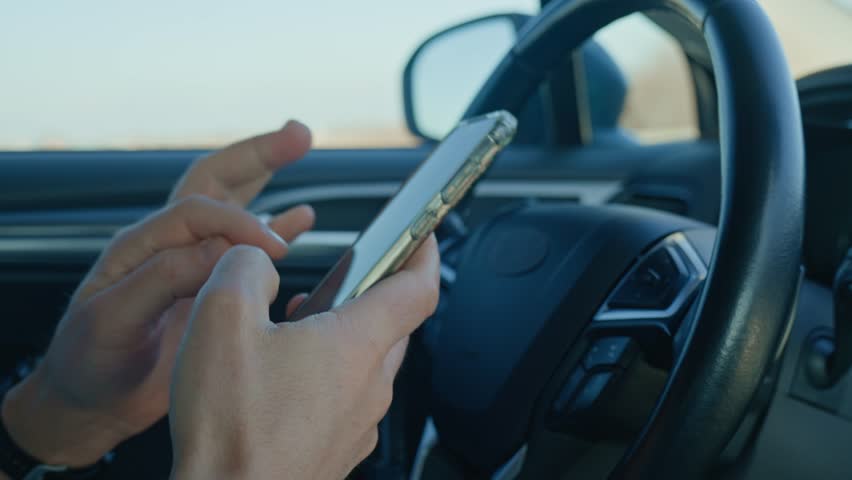 Close-up of driver hands using smartphone while driving car, risk of distraction and road safety concerns. Dangerous driving of vehicle, violation of traffic rules