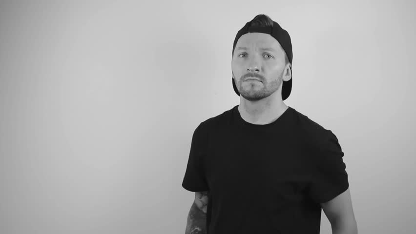 Black and white footage of a tattooed man with a sleeve crossing his arms and hands and nodding with approval and confidence in slow motion. Video is filmed on a white background that has shadows.
