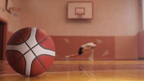 Basketball ball lies on wooden floor of gym with player stretching in background - Powered by Shutterstock - Get 15% off with code: PIKWIZARD15