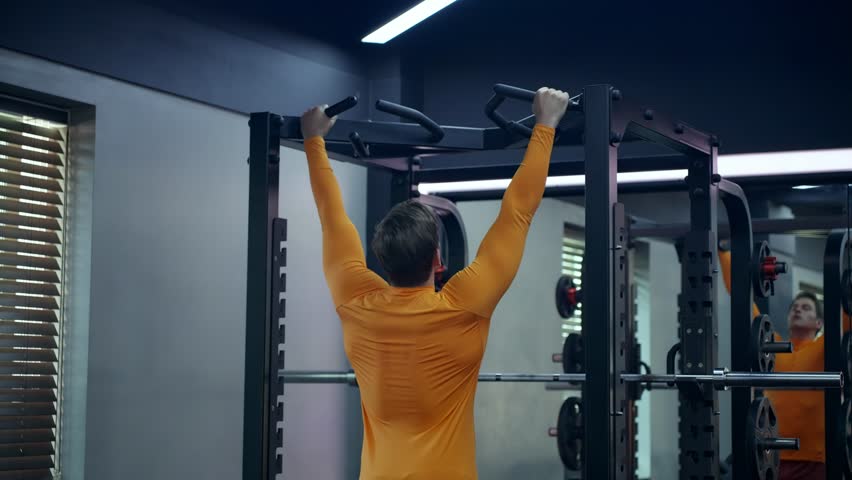 Crossfit athlete doing pull-ups at the gym - rear view. Handsome man in orange sportwear doing functional training on horizontal bar - slow motion