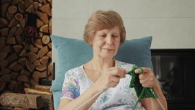 Elderly woman knitting with green yarn in a cozy armchair near a fireplace, concentrating on her craft with a calm and focused expression. High quality 4k footage - Powered by Shutterstock - Get 15% off with code: PIKWIZARD15