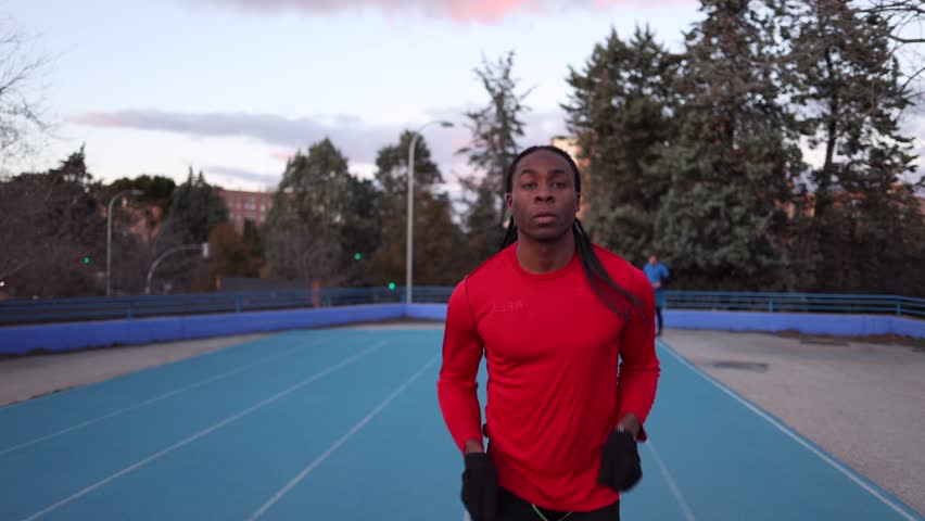 Determined young black athlete with long braids wearing a red long sleeved shirt and black leggings runs on a blue track in an urban park at sunset