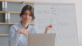 A young woman is leading an online educational session, using a laptop and a whiteboard at her home. She explains English verb conjugations, engaging her students remotely. - Powered by Shutterstock - Get 15% off with code: PIKWIZARD15