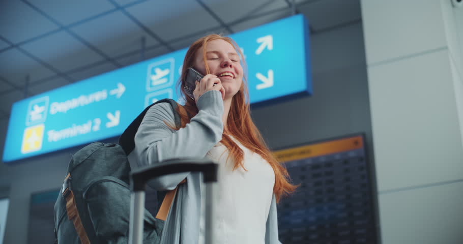 Crowded Airport Terminal: Caucasian Woman Holding Plane Ticket, Talking by Mobile Phone, Checking Flight Information. In the Background, Digital Arrival and Departure Display, Diverse People Walking.