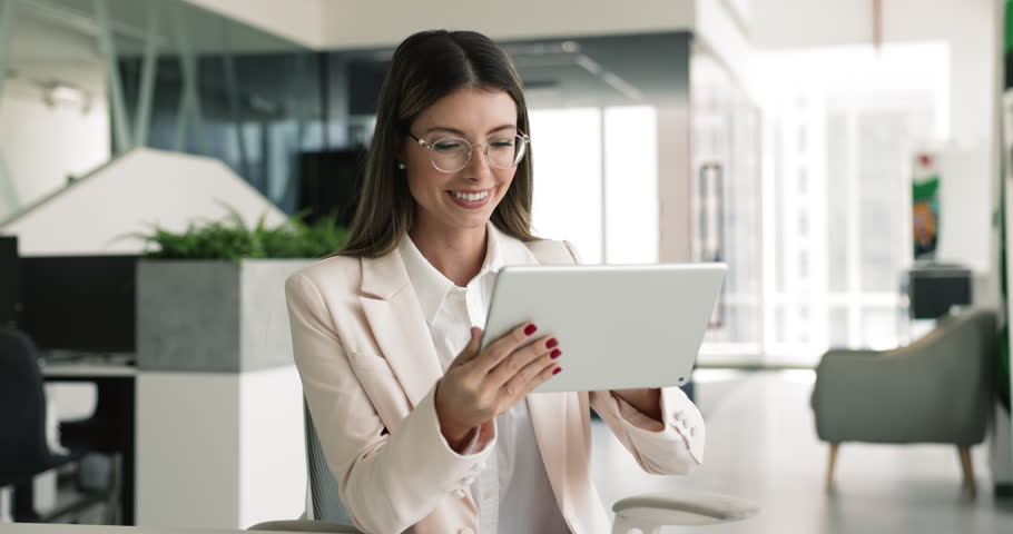 Focused confident young business professional woman in stylish glasses using tablet computer pad in office, working on project with gadget, checking Internet marketing statistic report, getting happy