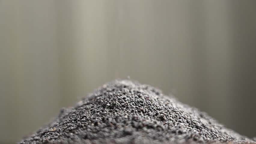 Fine poppy seeds falling onto a small pile in slow motion. Delicate grains forming a soft mound against a blurred background. Macro shot.