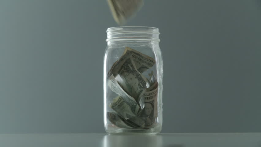 Hands put money into a tip jar or home savings, filling up the jar with dollars.