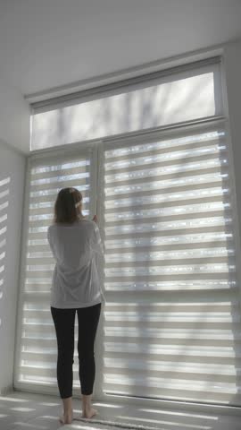 Vertical video of a Woman closing and opening modern room blinds with lighting range control. 4k footage.