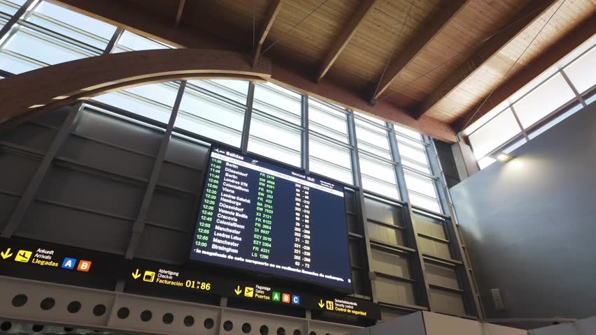A digital arrivals board displays incoming flights at a modern airport terminal. Sunlight streams through large windows, creating a bright, spacious atmosphere for travellers. (4K)