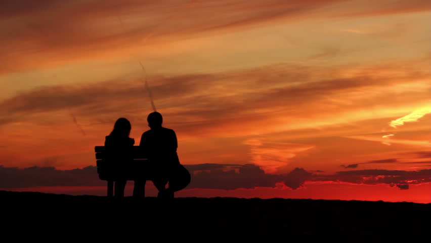 Silhouetted couple watch a beautiful sunset from a bench by the sea.