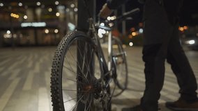 Male cyclist wearing protective mask parks his bicycle and straps it to pole outside bicycle parking lot on city evening in glow of city lights. Lack of bicycle infrastructure. Buckles up bicycle.  - Powered by Shutterstock - Get 15% off with code: PIKWIZARD15