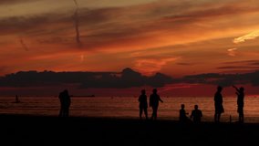 People silhouetted against a sunset sky, walking on the quayside next to the ocean. Clip ends with two people sitting alone.