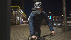 Man in helmet and medical mask rides his bike through city at night, parks it near streetlight, removes his helmet and hangs it on bikes handlebar, reflecting urban pollution and health concerns.  - Powered by Shutterstock - Get 15% off with code: PIKWIZARD15