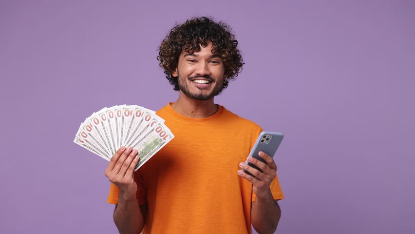 Happy joyful young Indian man he wear orange t-shirt casual clothes use mobile cell phone hold win fan of cash money in dollar banknotes isolated on plain purple background studio. Lifestyle concept