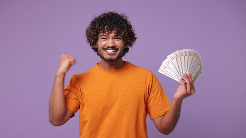 Happy young Indian man wears orange t-shirt casual clothes hold fan of cash money in dollar banknotes do winner gesture celebrate isolated on plain pastel light purple background. Lifestyle concept