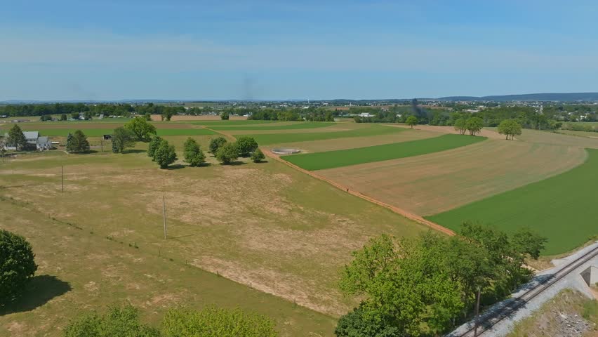 Expansive fields stretch across the landscape under a clear blue sky in Pennsylvania. Lush green areas contrast with dry land, illustrating the beauty of rural farming life.