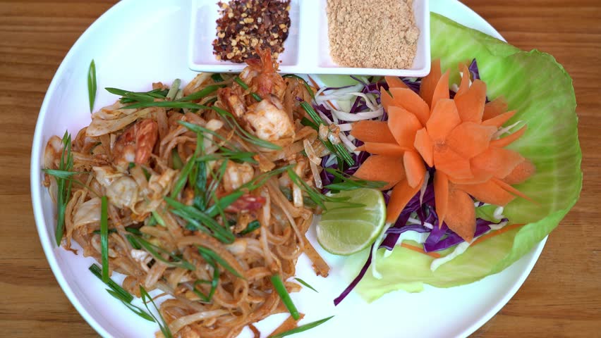 Traditional Thai pad thai with shrimp, rice noodles, green onions, and peanuts, served with a carved carrot garnish, cabbage salad, and lime wedge on a white plate. Concept of authentic Asian cuisine