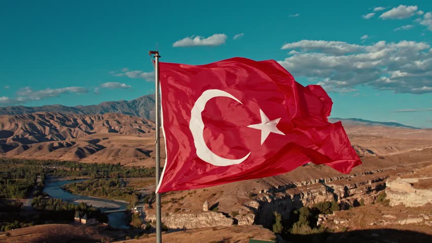 The Turkish flag waves in the wind against a breathtaking natural landscape. Mountains, a river, and the vast sky create a powerful and patriotic atmosphere.