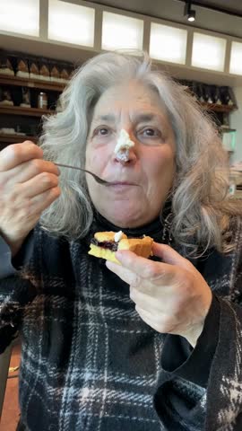 Senior woman savoring a sweet treat in a bakery, bringing a forkful of cream to her lips while holding a partially eaten pastry, creating a humorous moment of indulgence