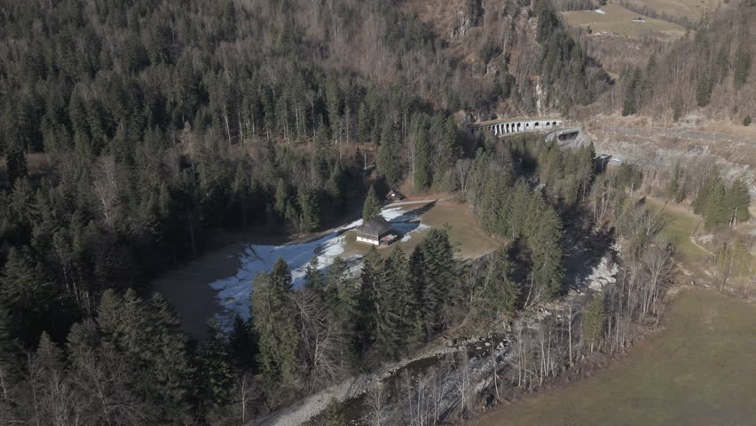 A scenic mountain road with a curved tunnel built into the rocky hillside, surrounded by a forest. Snow-capped peaks and alpine meadows are visible in the background, creating a stunning natural lands