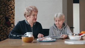 Two senior women sitting at a wooden table, discussing a recipe book while preparing ingredients for cooking in a warm and cozy home setting. High quality 4k footage - Powered by Shutterstock - Get 15% off with code: PIKWIZARD15