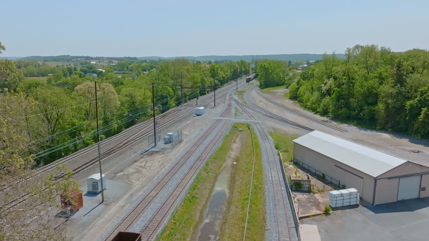 The serene landscape features intersecting train tracks surrounded by vibrant green trees and open fields. A few trains pass near the junction, of a new freight yard, creating a lively atmosphere.