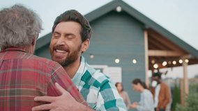 Camera focus on Caucasian man in checkered shirt coming up to mature father. Men hugging each other while smiling. Family reunion. Son visiting dad in village. Lights shining on house in background. - Powered by Shutterstock - Get 15% off with code: PIKWIZARD15